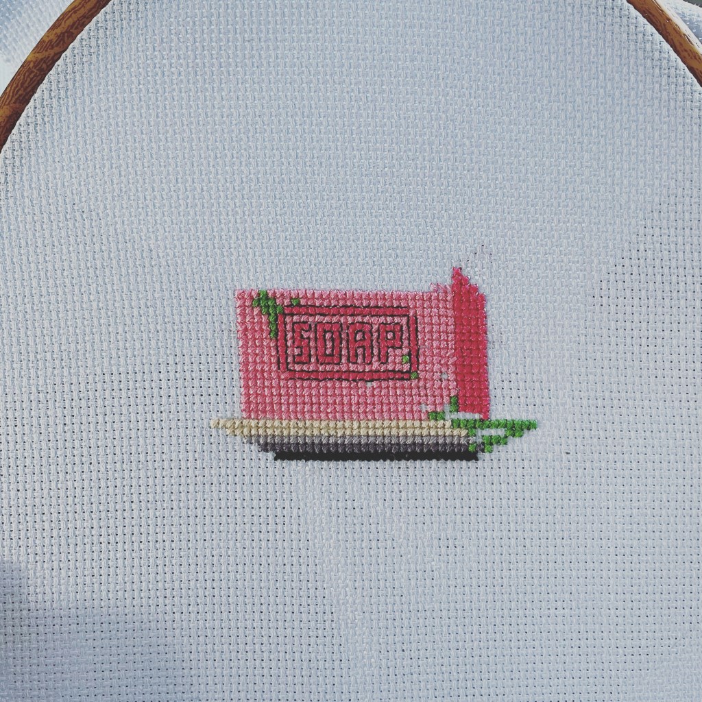 a blue cross stitch fabric, with a pink soap bar sitting on a soap dish. The soap has green slime in the corner