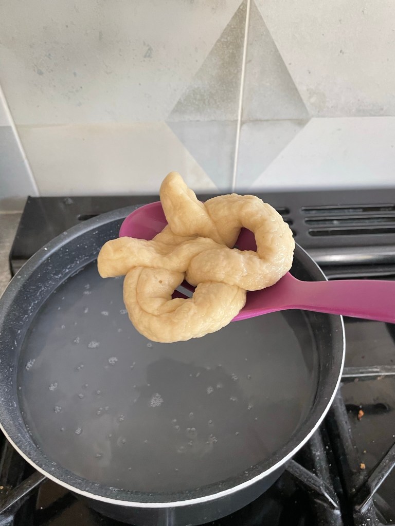 a dough shaped like a pretzel on a purple spoon above a pan of hot water.